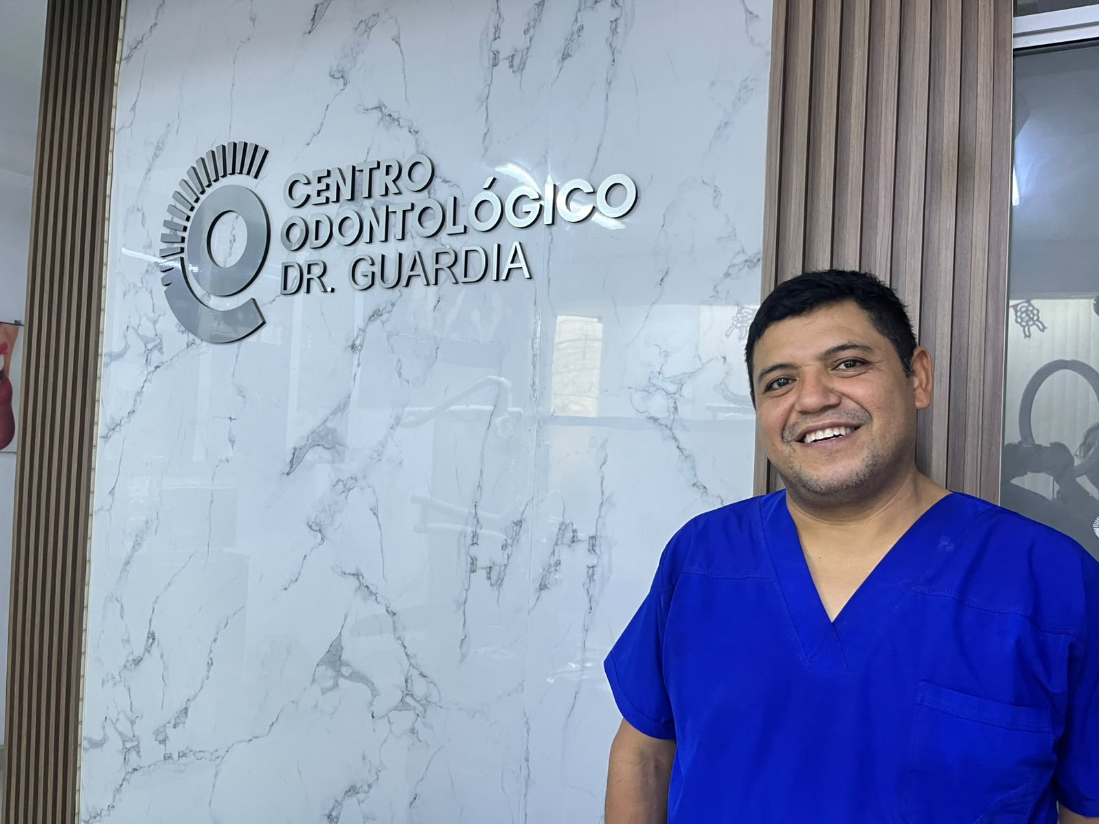 Dr. Guardia sonriendo, con uniforme clínico azul, en el consultorio
